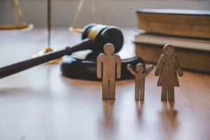 Wooden cutouts representing a father, child with raised arms, and mother standing in the foreground on a wooden desk, with a judge's gavel, law books, and brass scales of justice resting blurred in the background.