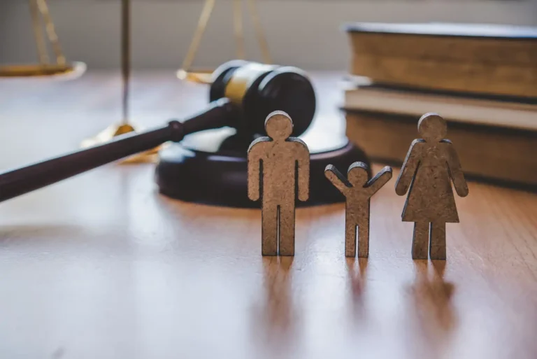 Wooden cutouts representing a father, child with raised arms, and mother standing in the foreground on a wooden desk, with a judge's gavel, law books, and brass scales of justice resting blurred in the background.